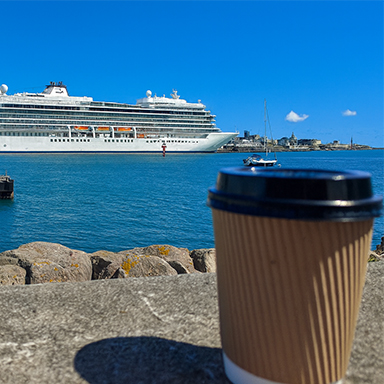 A large cruise ship docked in a harbor with a distinctive tower in the background and a takeaway coffee cup in the foreground.