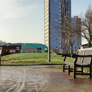 A bench on a brick walkway with a building and The Daily Grind coffee van visible in the background, creating a serene outdoor setting.