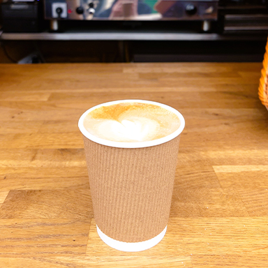 A paper cup of cappuccino with foam sits on a wooden counter in front of a professional espresso machine in a coffee shop.