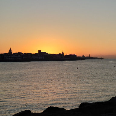 Sunset casts vibrant colors over a port, highlighting boats and the skyline against an sky.